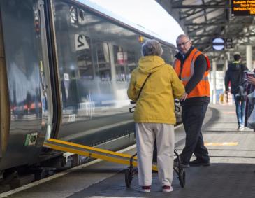 Staff member assisting passenger with mobility aid onto train with ramp deployed at a train station